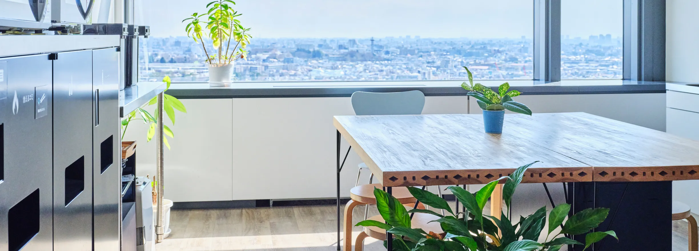 大きな窓から街を一望できる明るいオフィスの休憩スペース。木製のテーブルと観葉植物が配置され、自然光が差し込む心地よい空間