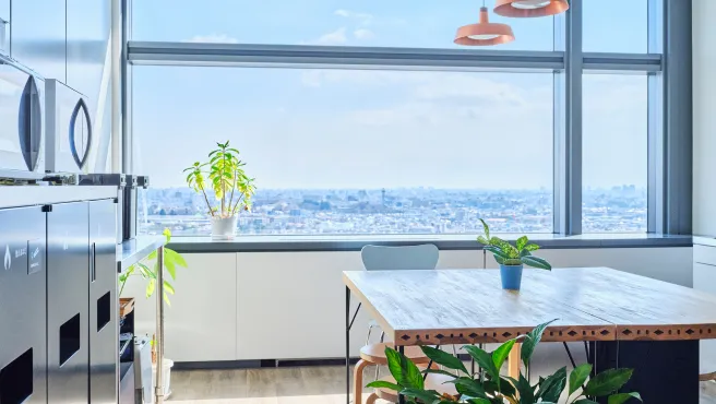 A bright office break space with a panoramic view of the city from a large window. Wooden tables and potted plants create a comfortable space filled with natural light.