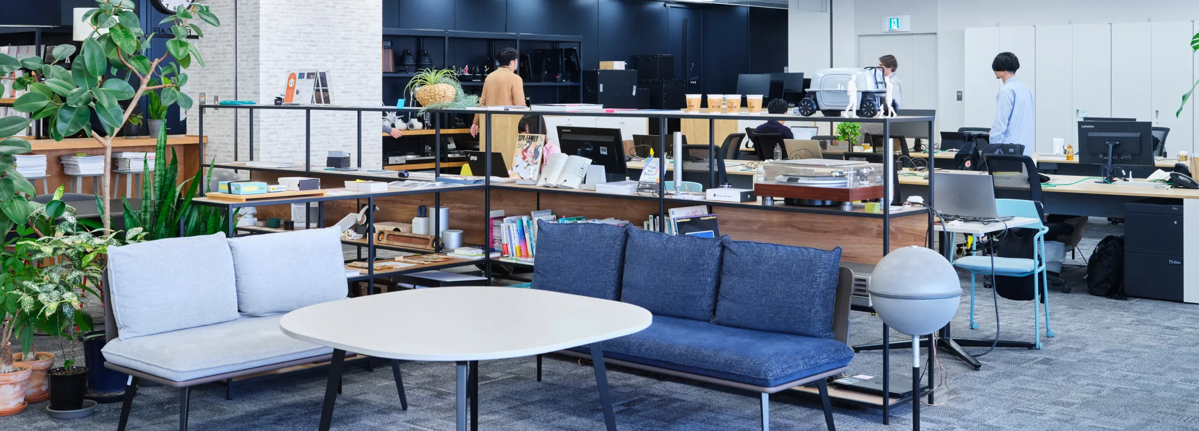 A relaxing space in an open office, lined with potted plants and books. There is a sofa and table in the foreground, and employees working in the background.