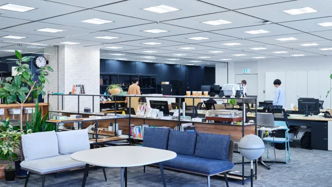 A relaxing space in an open office, lined with potted plants and books. There is a sofa and table in the foreground, and employees working in the background.