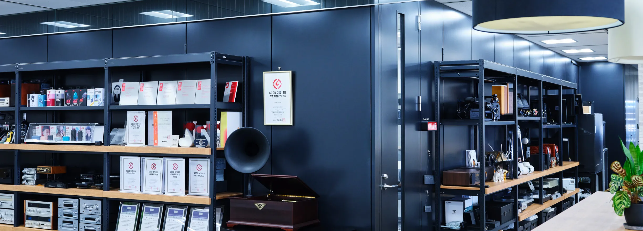 An exhibition space in a corner of the office. Audio equipment, headphones, design award trophies, and certificates are neatly arranged on black metal racks. A certificate for the "GOOD DESIGN AWARD 2023" is displayed on the wall, and a classic gramophone is also on display.