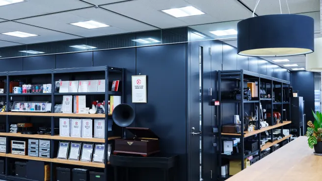 An exhibition space in a corner of the office. Audio equipment, headphones, design award trophies, and certificates are neatly arranged on black metal racks. A certificate for the "GOOD DESIGN AWARD 2023" is displayed on the wall, and a classic gramophone is also on display.